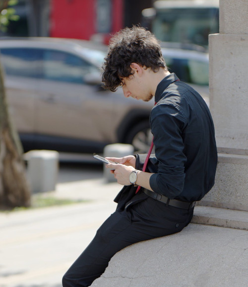 A young man sitting outside texting on his phone